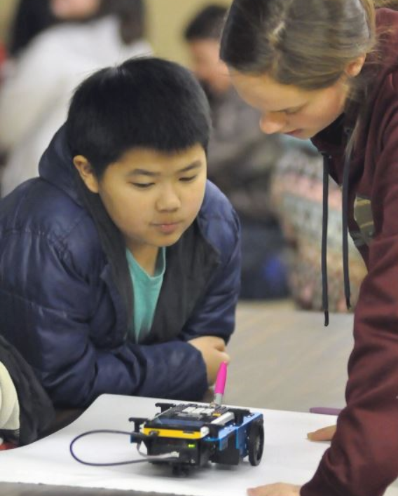 Keota third grade student Kenjie Her watches with fascination as junior Sierra Bishop helps draw a robot using the TI-Innovator Rover.